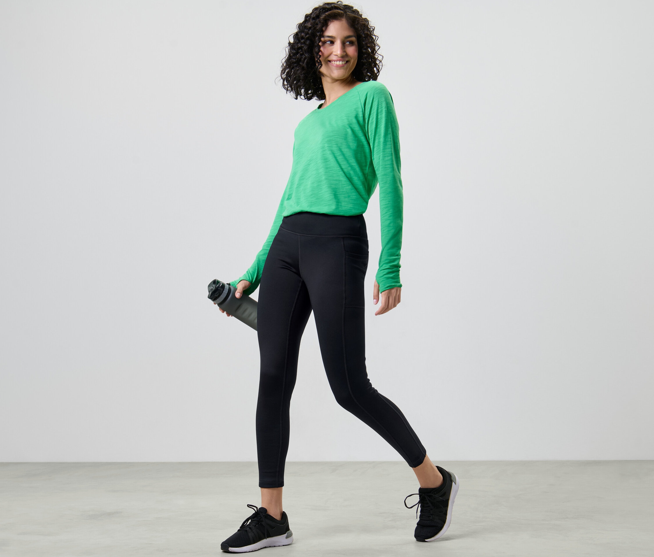 Žena v zeleném tričku a termo legínách drží láhev s vodou.