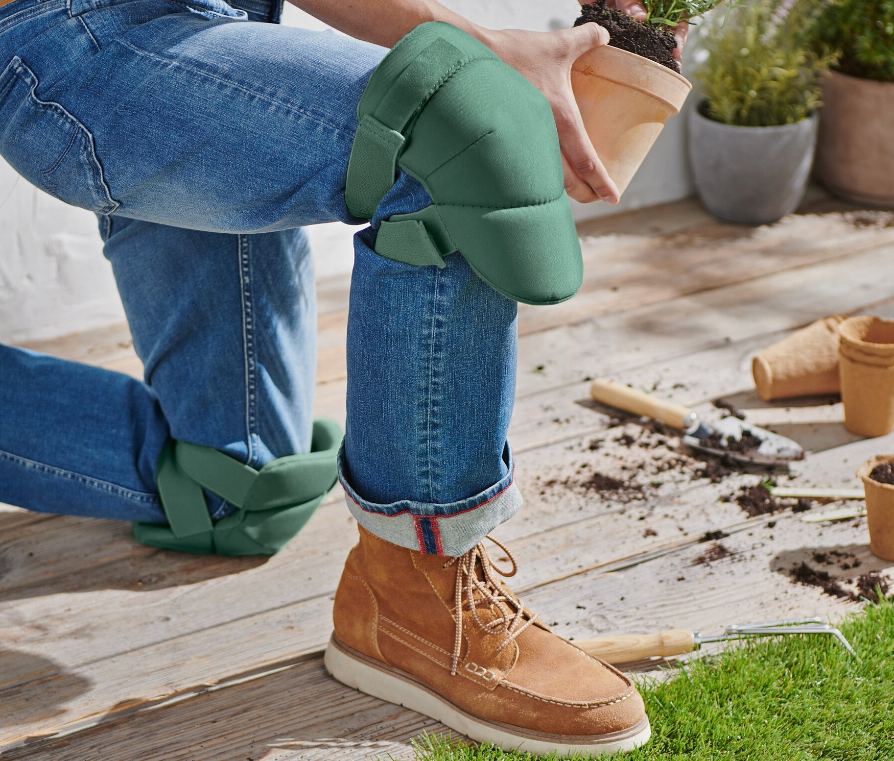Osoba na kolenou s nasazenými zahradními chrániči kolen drží květináč s rostlinou.