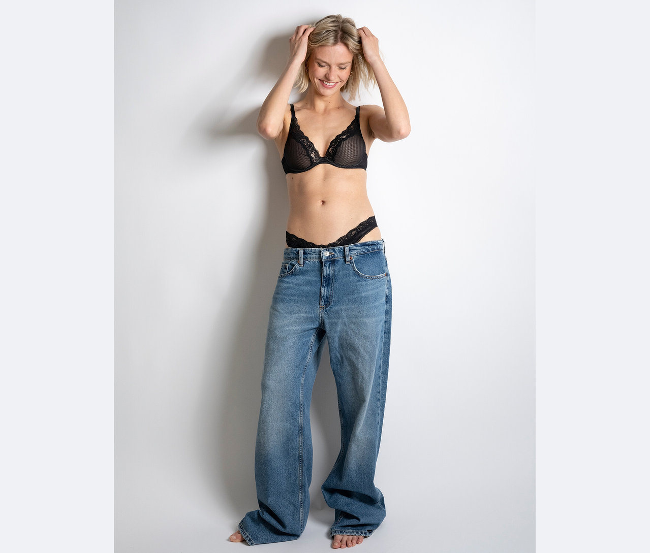 Žena se usmívá a drží si ruce na hlavě, má na sobě černou krajkovou podprsenku a tanga a obrovské džíny.