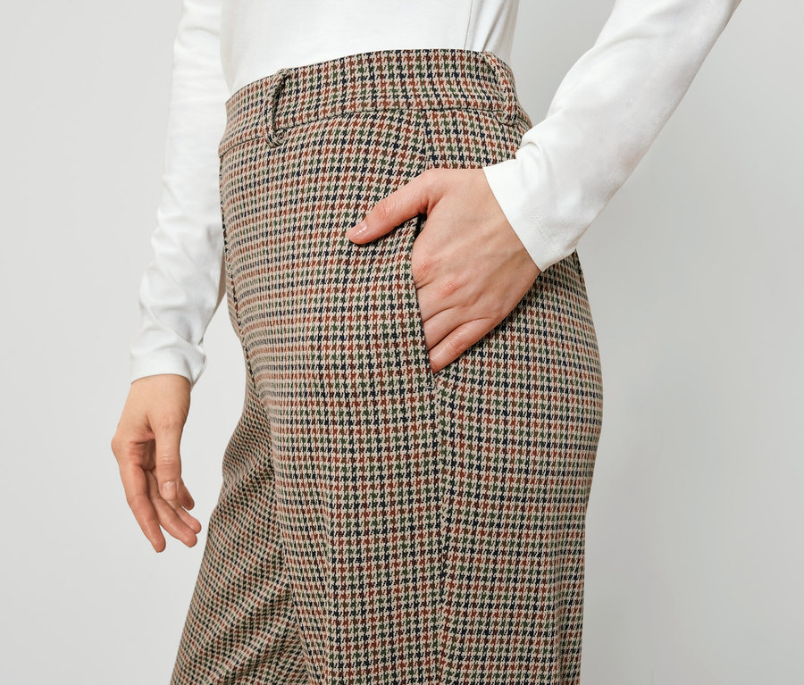 Žena má ruku v kapse hnědých kostkovaných kalhot.