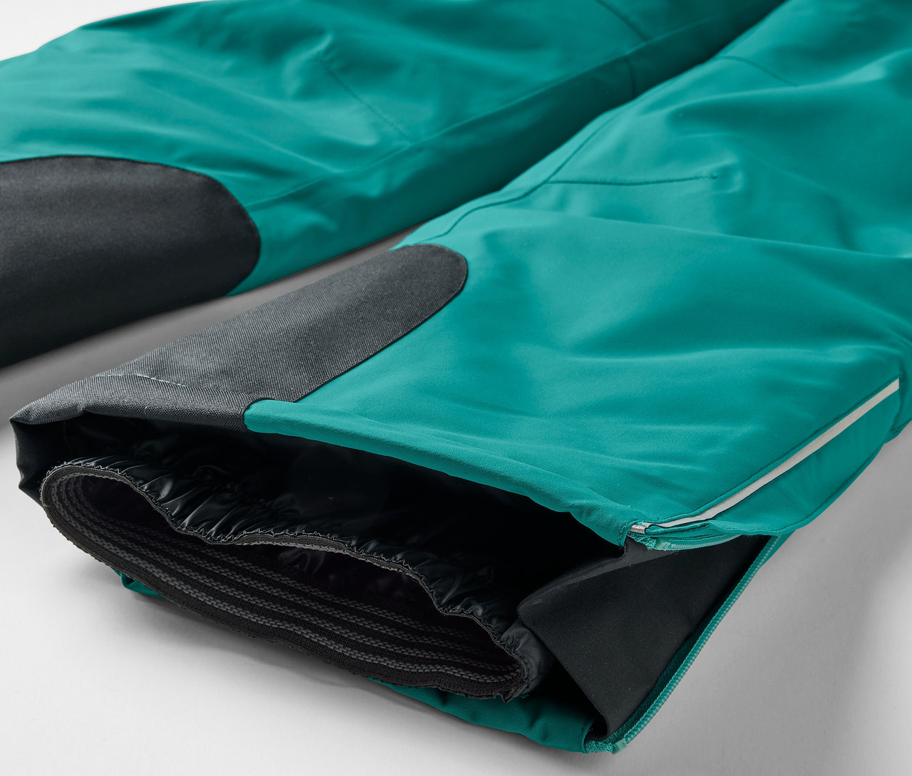 Detailní pohled na turkysové kalhoty s černými nášivkami na kolenou a rozepnutým lemem nohavice odhalujícím černou podšívku.