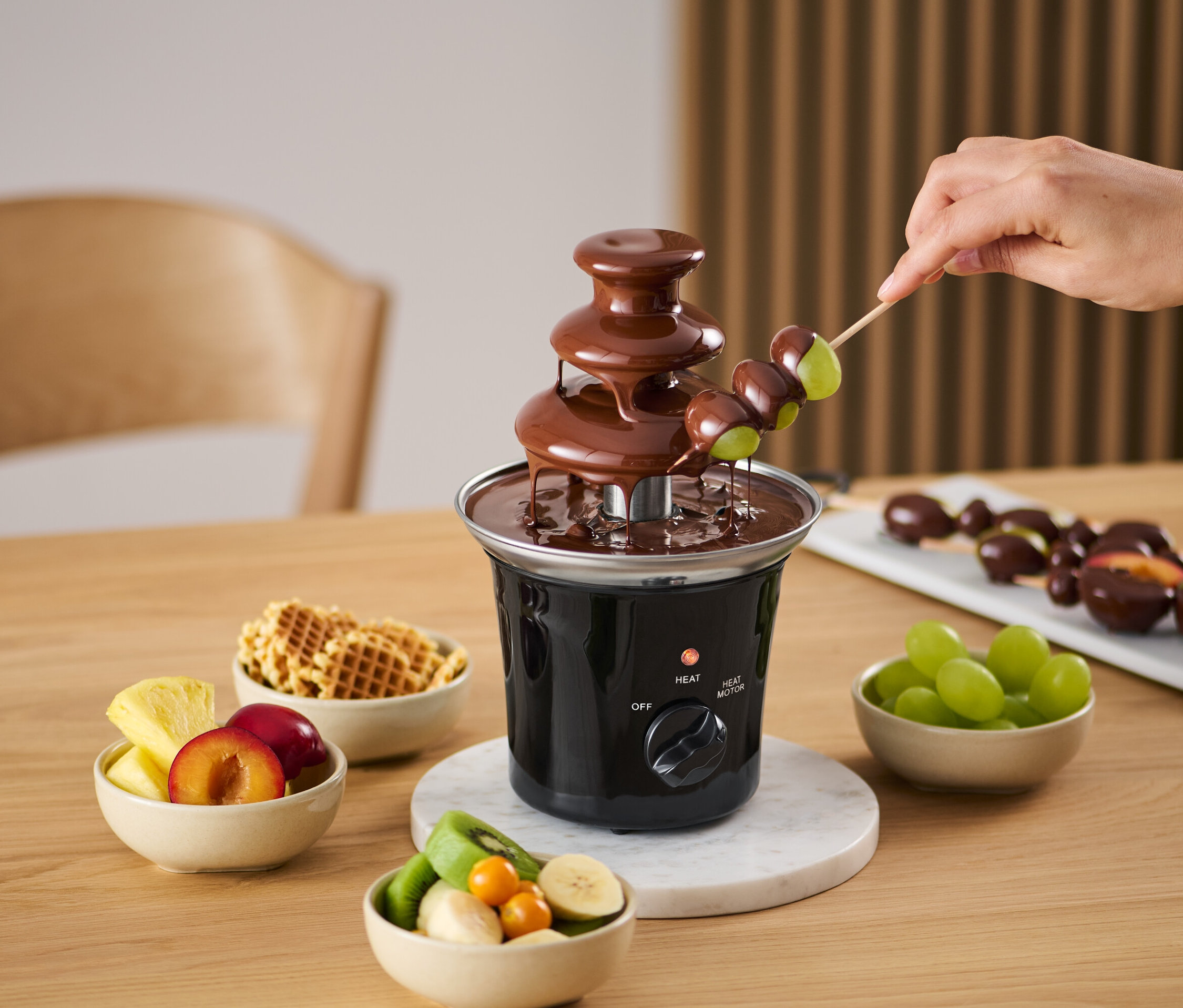 Ruka namáčí hrozny na špejli v čokoládové fontáně. Okolo jsou misky s ovocem a vaflemi.