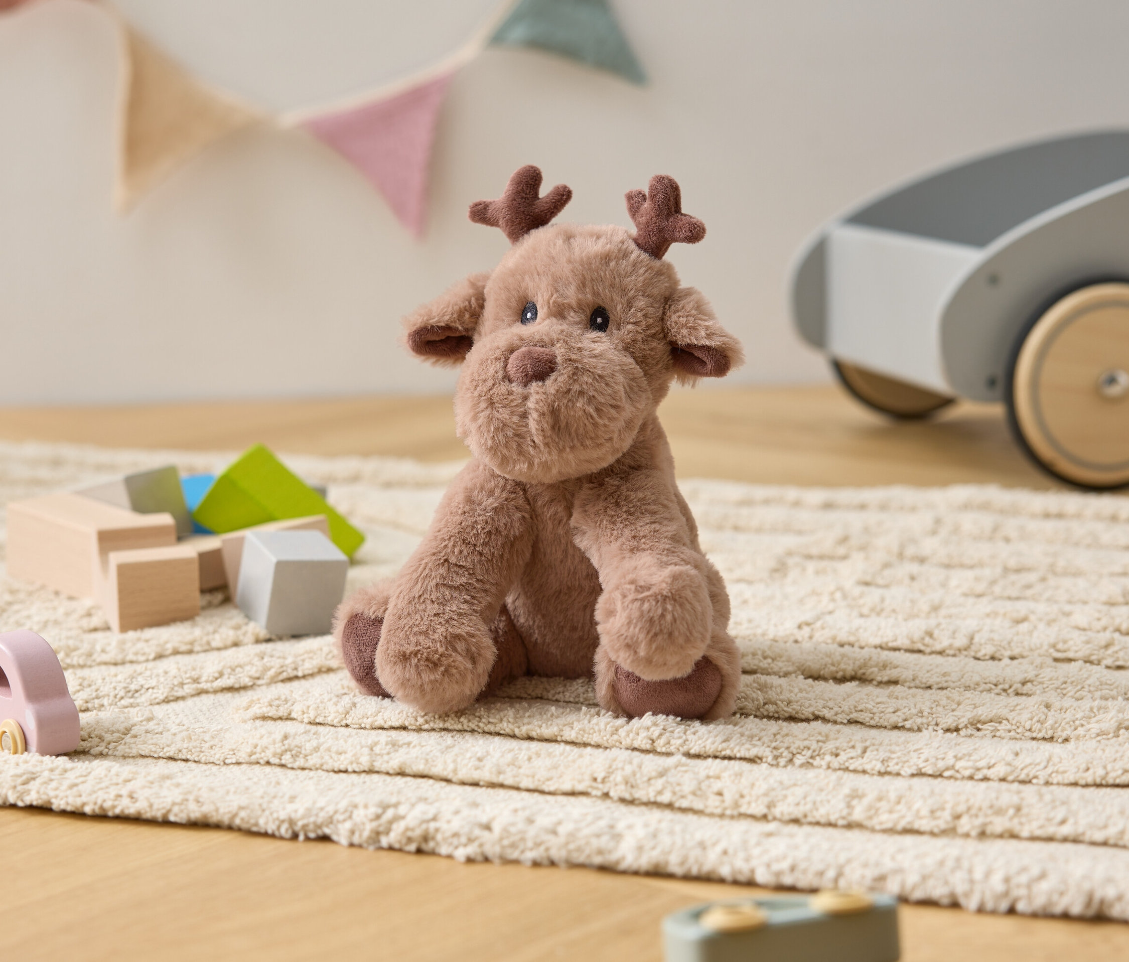 Plyšový los sedí na krémovém koberci mezi dřevěnými hračkami a šedým vozíkem.