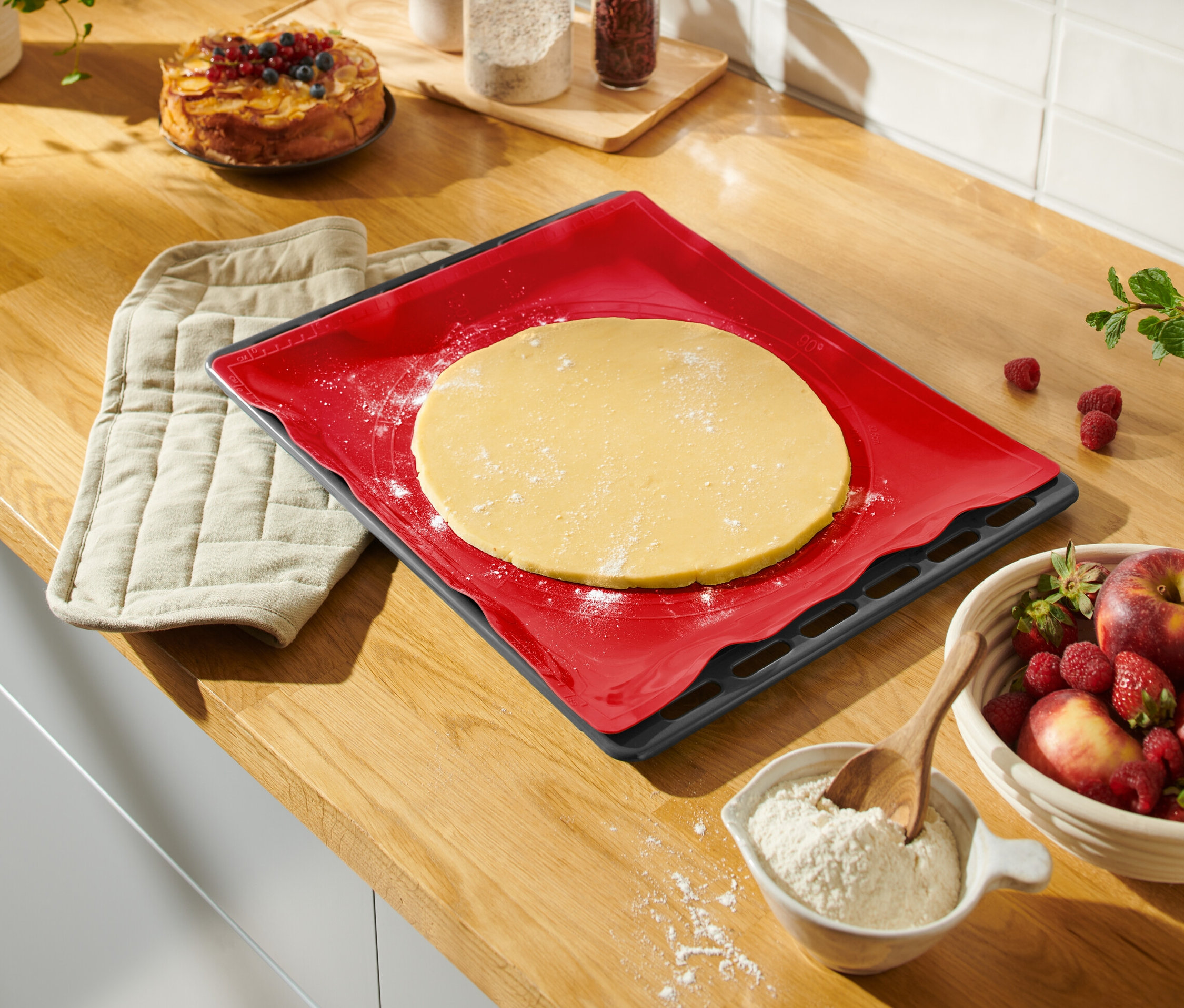 Na lince leží červená podložka na pečení s rozváleným těstem, vedle je chňapka, mísa s ovocem a koláč.