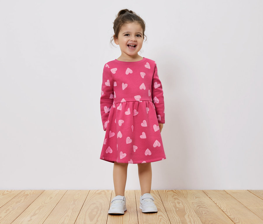 Usmívající se holčička stojí na dřevěné podlaze a má na sobě růžové šaty s dlouhým rukávem a potiskem srdíček.