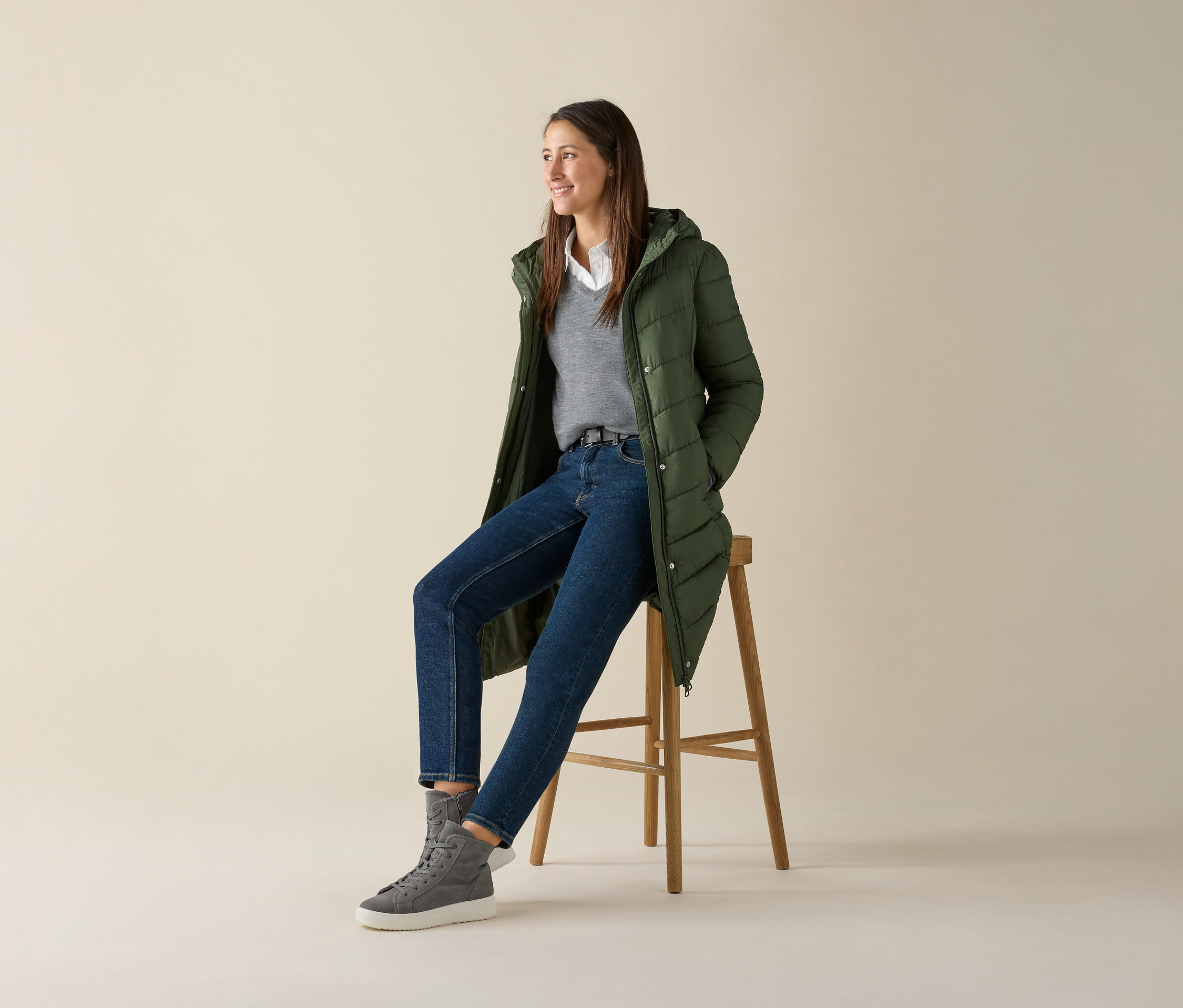Žena sedí na stoličce a má na sobě šedý svetr z jemného úpletu merino, džíny a zelenou bundu.