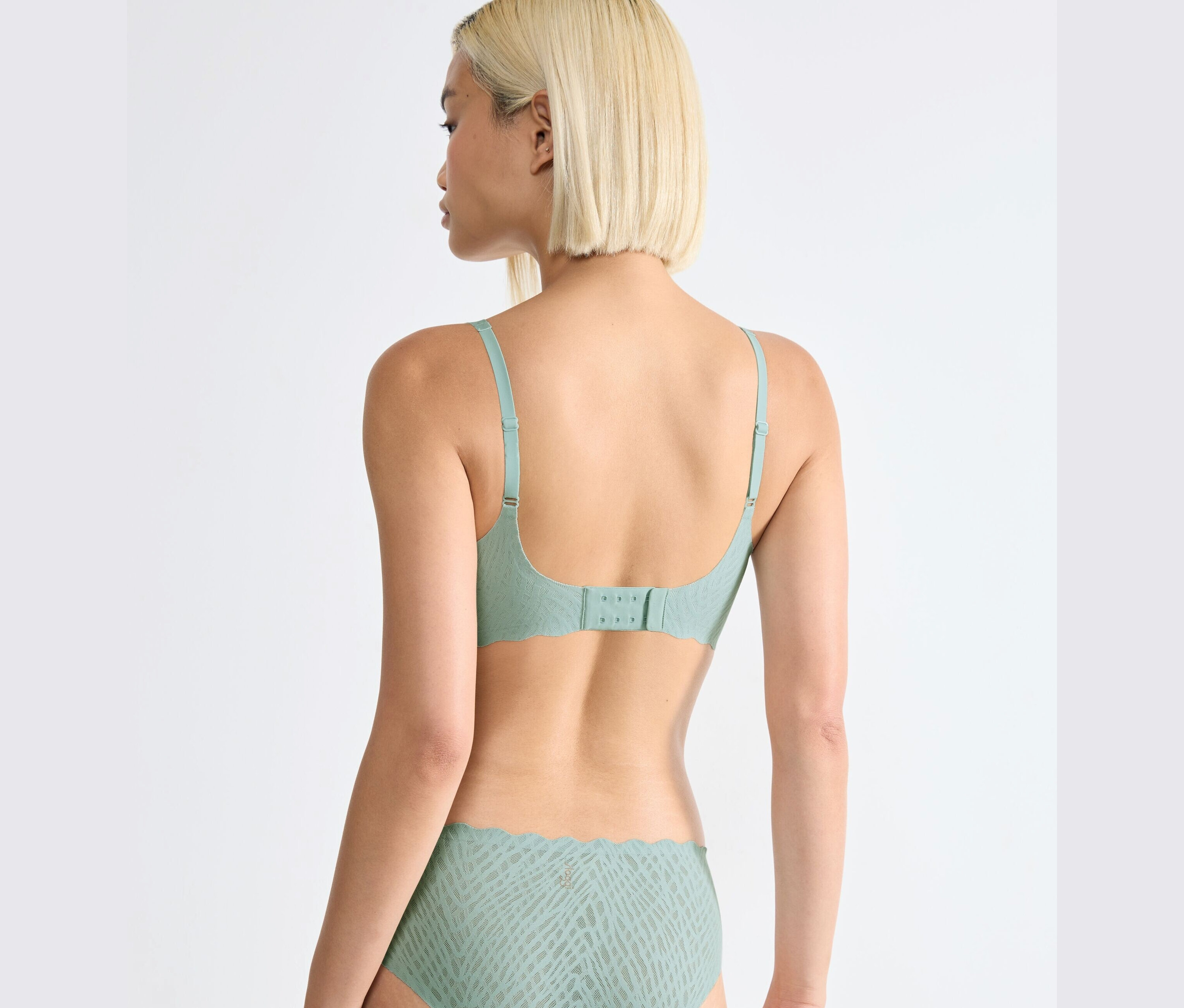 Pohled zezadu na ženu s blond vlasy v soupravě spodního prádla: Měkká podprsenka sloggi ZERO Feel Bliss Soft Bra, v barvě misty turquoise a odpovídající kalhotky.