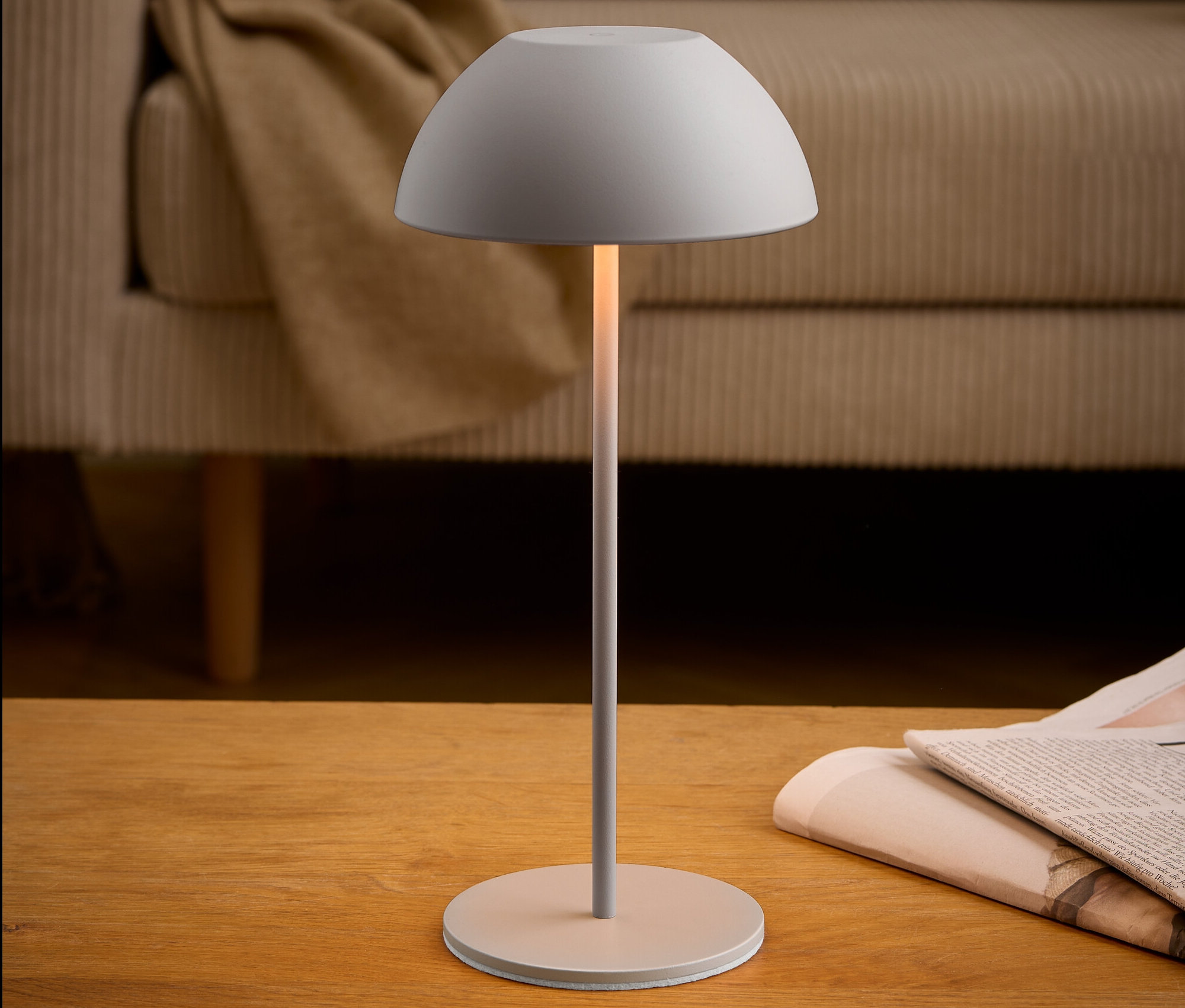 Šedá lampa stojí na dřevěném stole vedle novin. V pozadí je béžová pohovka.