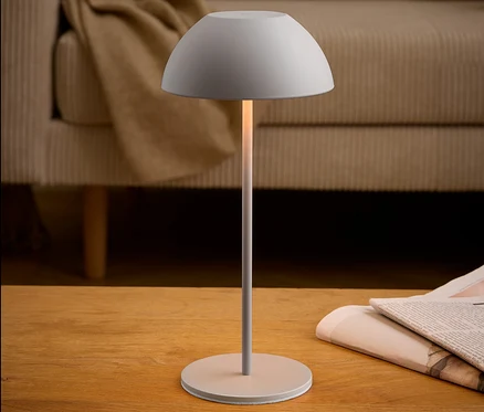 Šedá lampa stojí na dřevěném stole vedle novin. V pozadí je béžová pohovka.