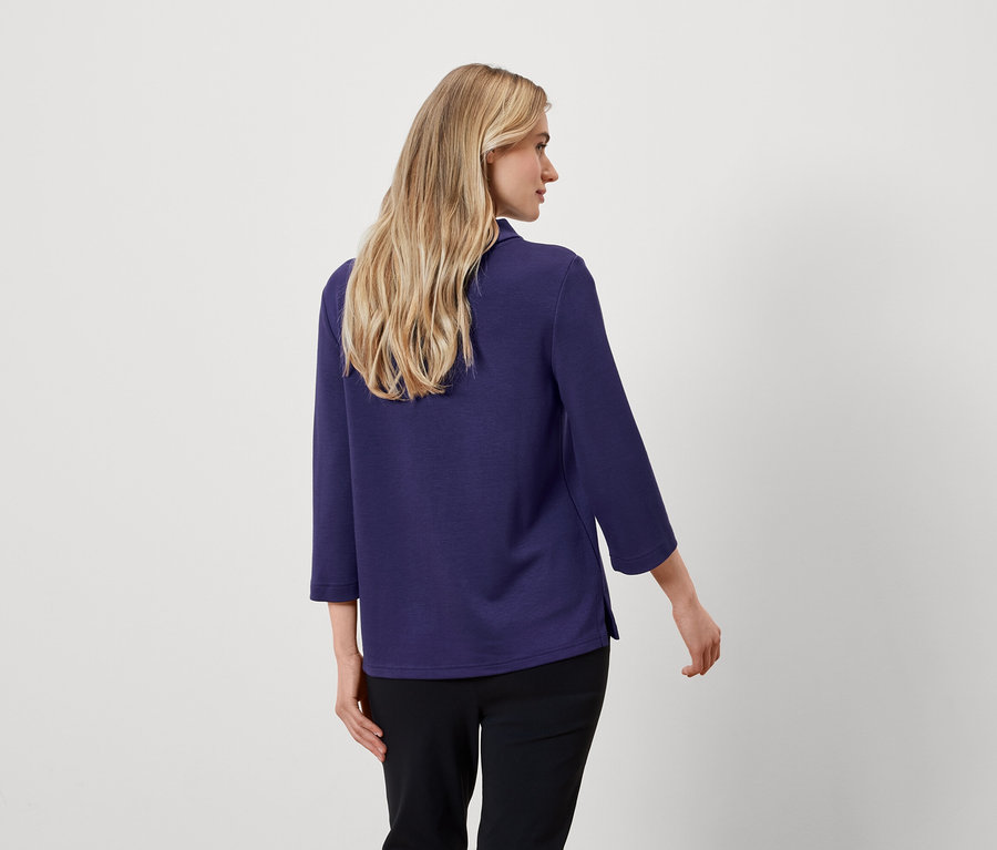 Zezadu vidíme ženu s blond vlasy, oblečenou v modré halence s tříčtvrtečními rukávy a černých kalhotách.