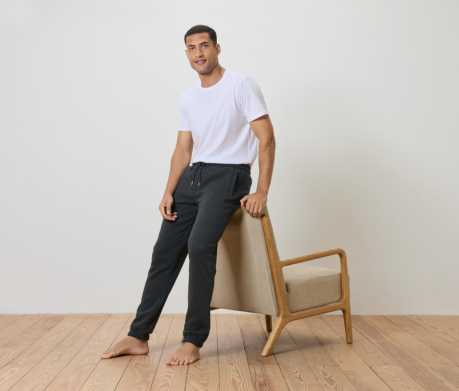 Muž opřený o židli, má na sobě bílé tričko a šedé tepláky.