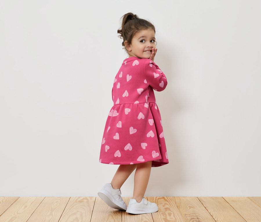 Dívka v růžových šatech se srdíčky a bílých teniskách se dívá přes rameno.