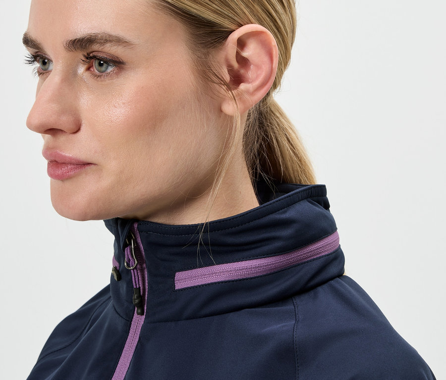 Detail obličeje ženy s blond vlasy staženými do culíku, oblečené do modré bundy s fialovými detaily.