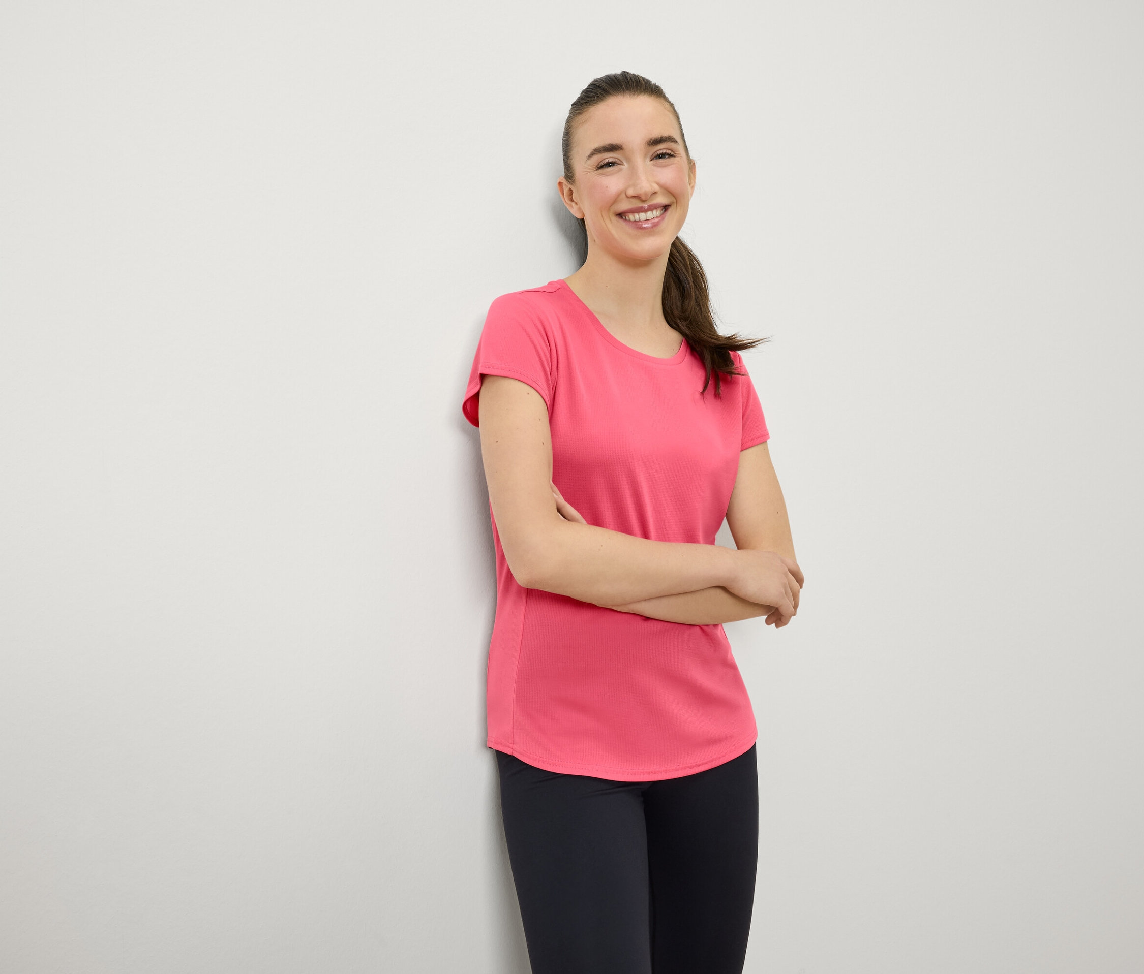 Usmívající se žena s hnědými vlasy v culíku stojí s rukama zkříženýma na hrudi. Má na sobě růžové tričko a tmavě modré legíny.