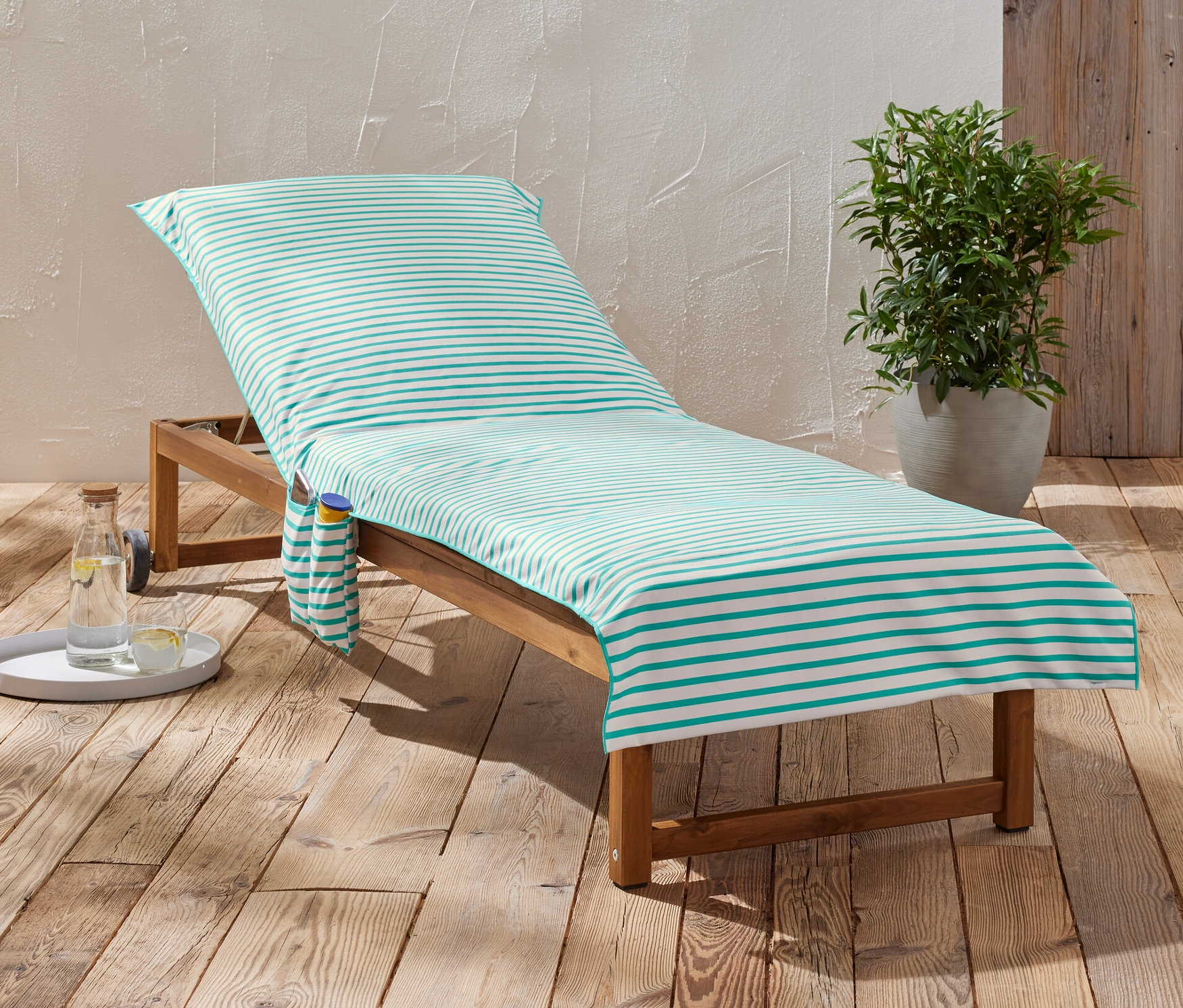 Pruhovaná venkovní osuška leží na dřevěném lehátku vedle nápoje a rostliny.