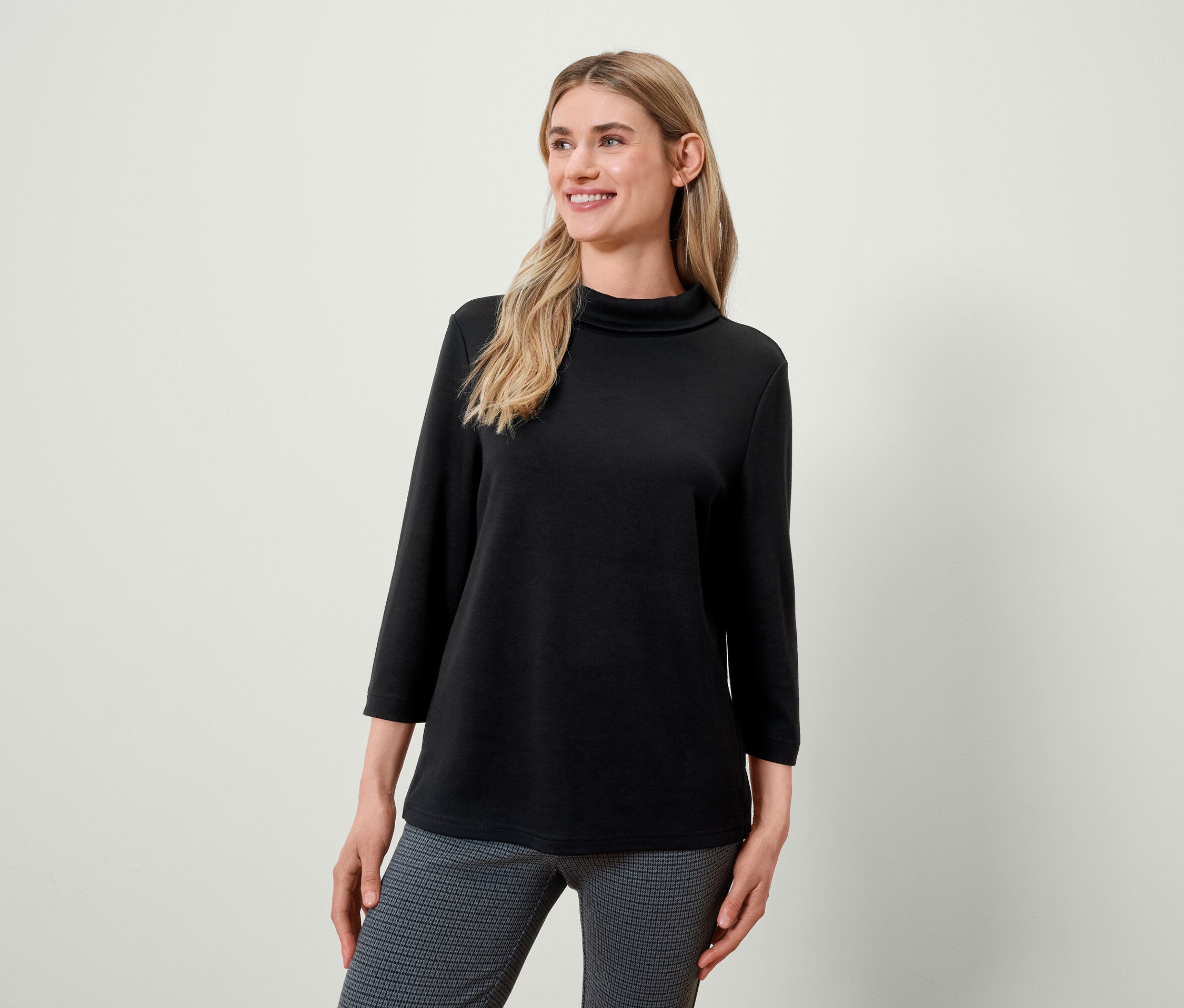 Blondýna s černým rolákem a šedými vzorovanými kalhotami se usmívá a dívá se doleva.