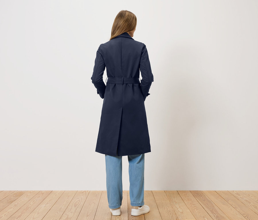 Žena stojí zády ke kameře a má na sobě tmavě modrý trenčkot, džíny a bílé boty.