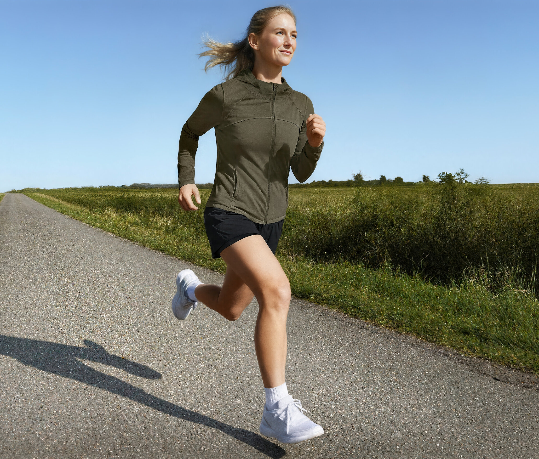 Žena běží po venkovské silnici ve sportovní bundě a běžeckých šortkách 2 v 1.