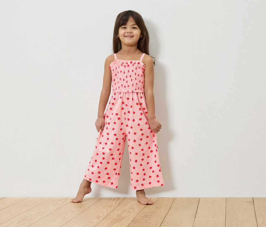 Dívka stojí bosá před bílou zdí a má na sobě růžový overal s jahodovým vzorem.