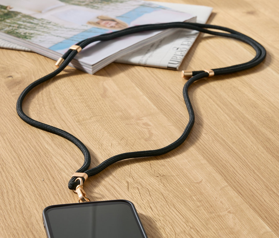 Černý řetízek na mobil s detaily ve zlaté barvě leží na stole vedle časopisu a béžové kabelky.