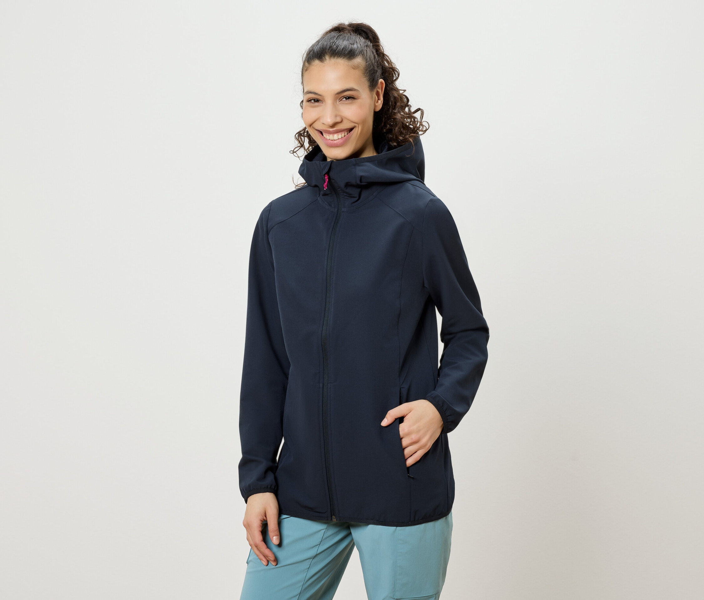 Usmívající se žena s kudrnatými vlasy sepnutými do culíku, oblečená do tmavě modré bundy s kapucí a modrých kalhot, s rukou v kapse bundy.