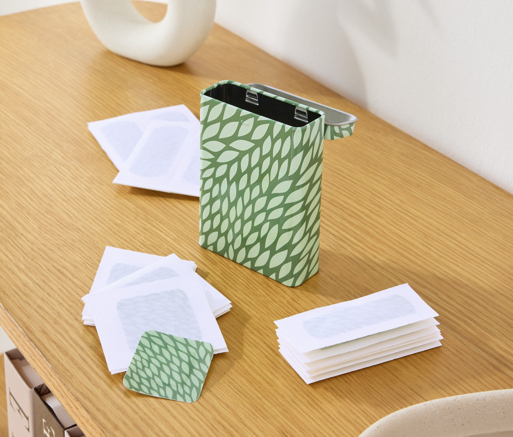 Na dřevěném stole stojí plechovka se zeleným vzorem listů, prázdné voskové proužky a váza s rostlinami.