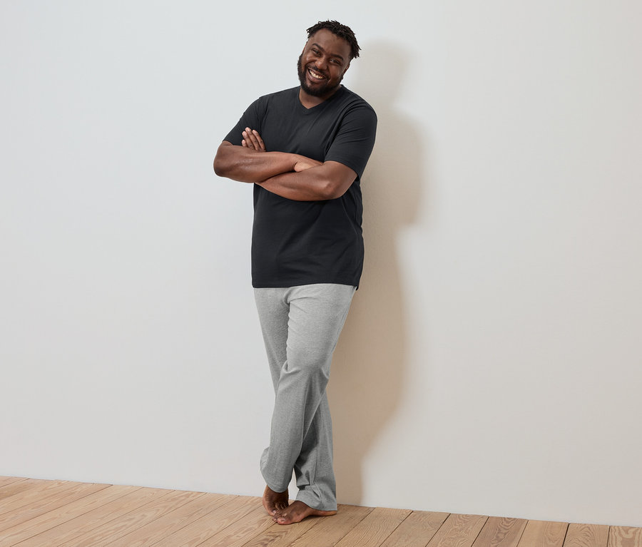 Tmavý muž se zkříženýma rukama se opírá o zeď, má na sobě černé tričko a šedé kalhoty.