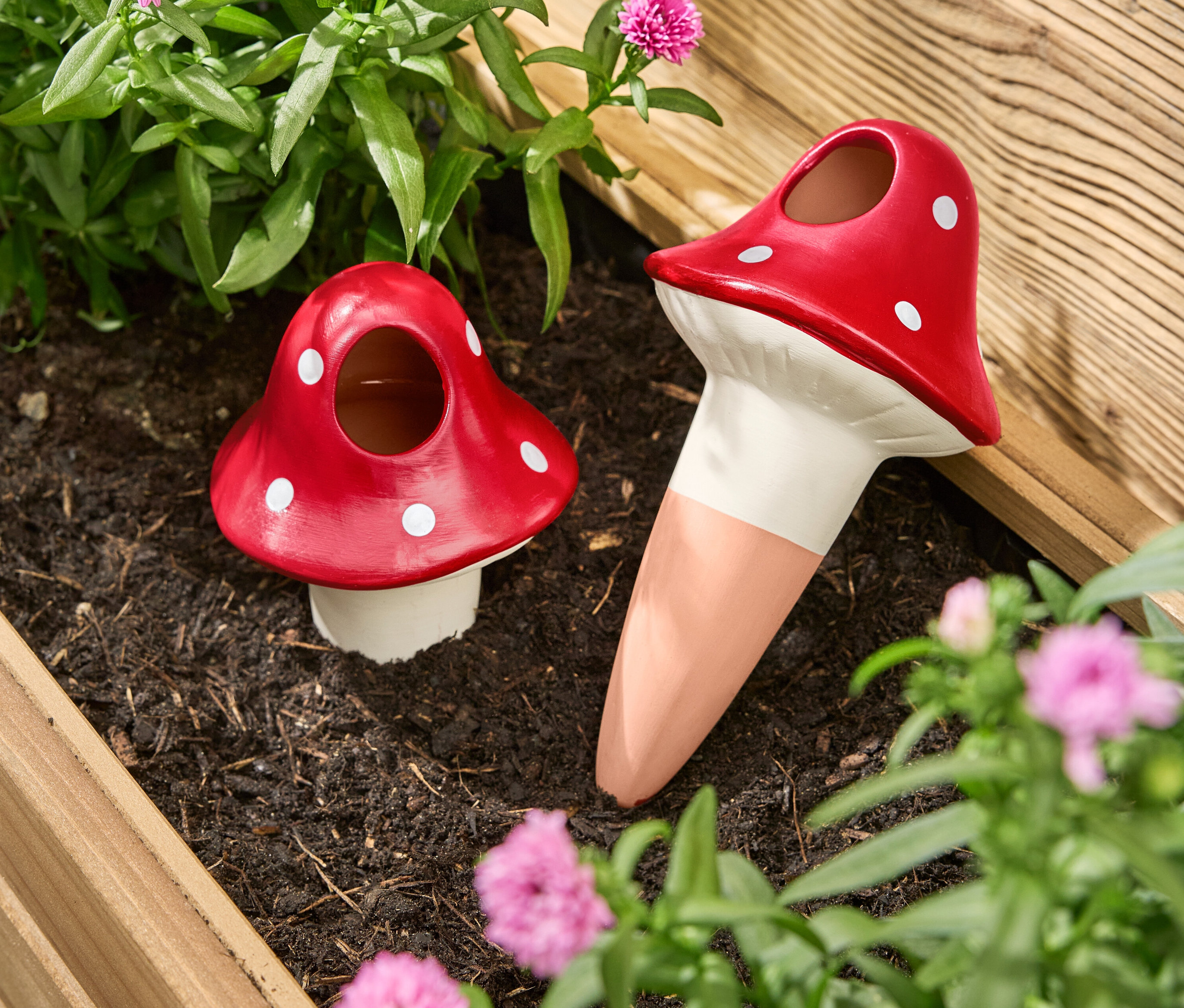 Dvě červené zavlažovací koule ve tvaru houby s bílými tečkami stojí v truhlíku mezi květinami a rostlinami.