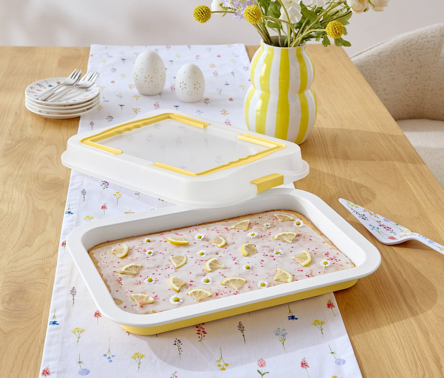 Na dřevěném stole leží dort s citronovou polevou a sedmikráskami, žluto-bílý pruhovaný džbán, lopatka na dort s květinovým vzorem a bílý ubrus s květinovým vzorem.