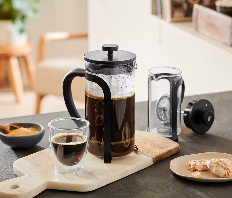 Černá konvička french press 800 ml, 6 šálků, naplněná kávou, stojí na mramorovém prkénku vedle sklenice kávy, misky s cukrem a sušenek. Vedle je rozebraná další konvička french press.