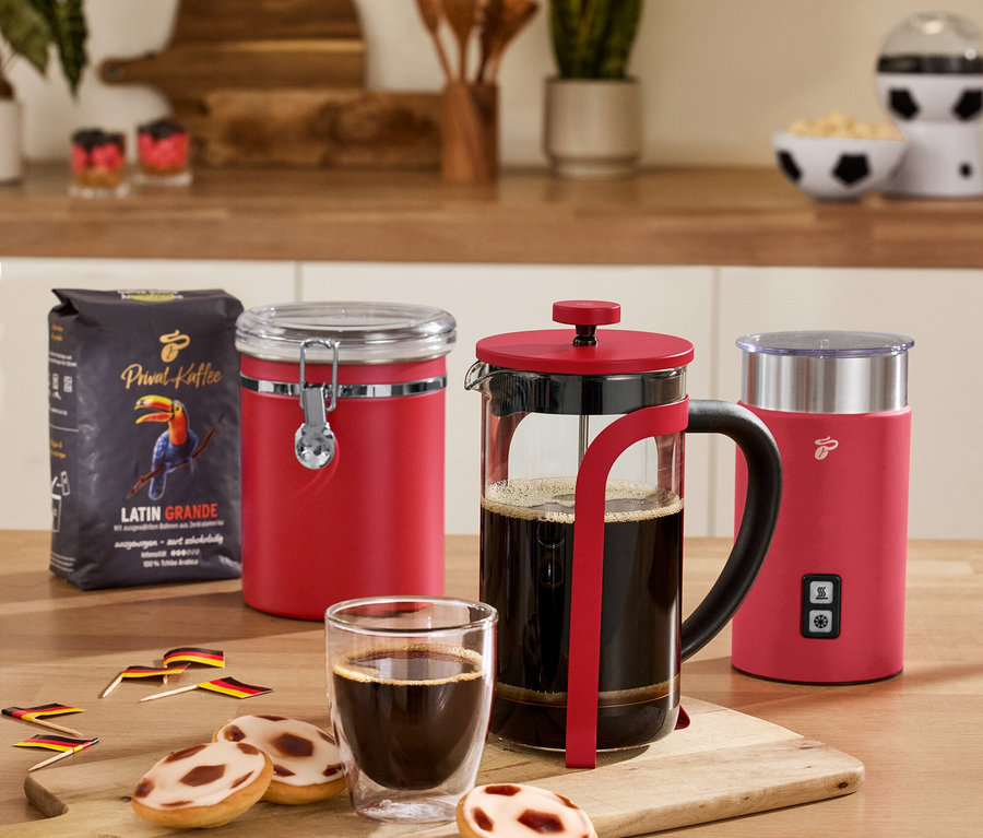Na stole stojí červená vzduchotěsná dóza na kávu, červený elektrický napěňovač mléka, červená konvička french press, 2 sklenice Caffè Crema a balíček kávy.
