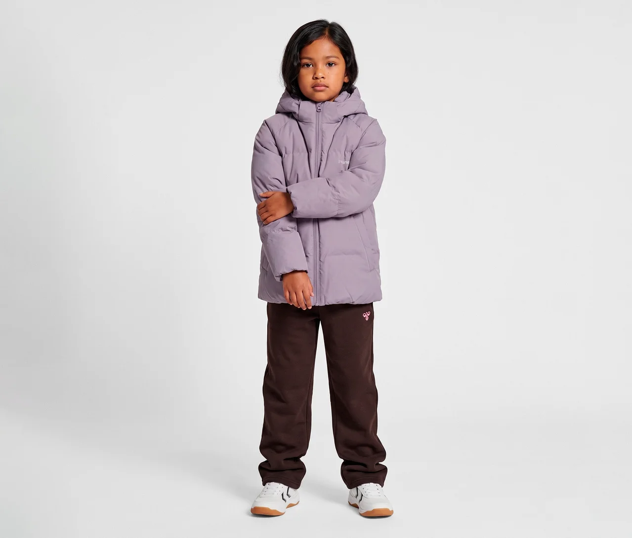 Dítě s tmavými vlasy stojí čelně, oblečené ve světle fialové bundě hummel® »HMLJR All Weather Puffer Jacket« s kapucí, tmavě hnědých teplácích a bílých teniskách. Ruce dítěte jsou zkřížené.