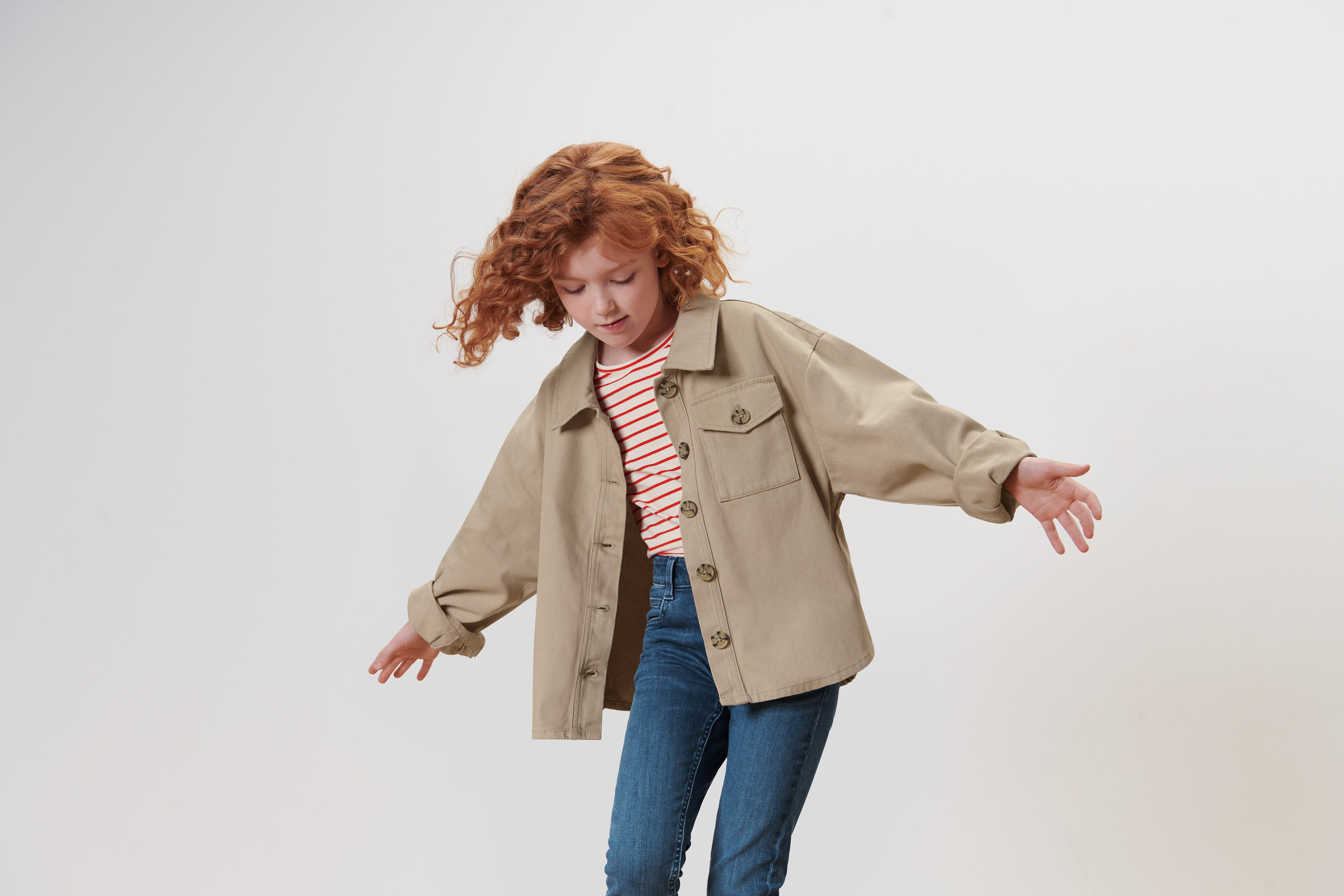 Dívka s rusými kudrnatými vlasy mává rukama a má na sobě džínovou bundu.