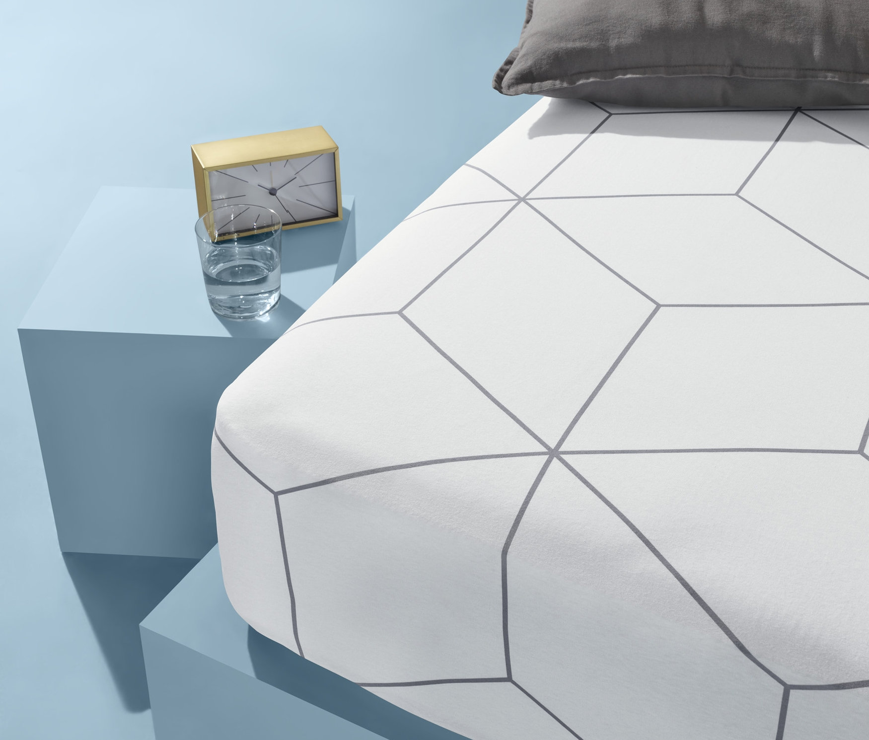 Na modrém pozadí stojí postel s bílým povlečením s geometrickým vzorem a šedým polštářem. Vedle postele na modrém stolku stojí zlaté hodiny a sklenice vody.