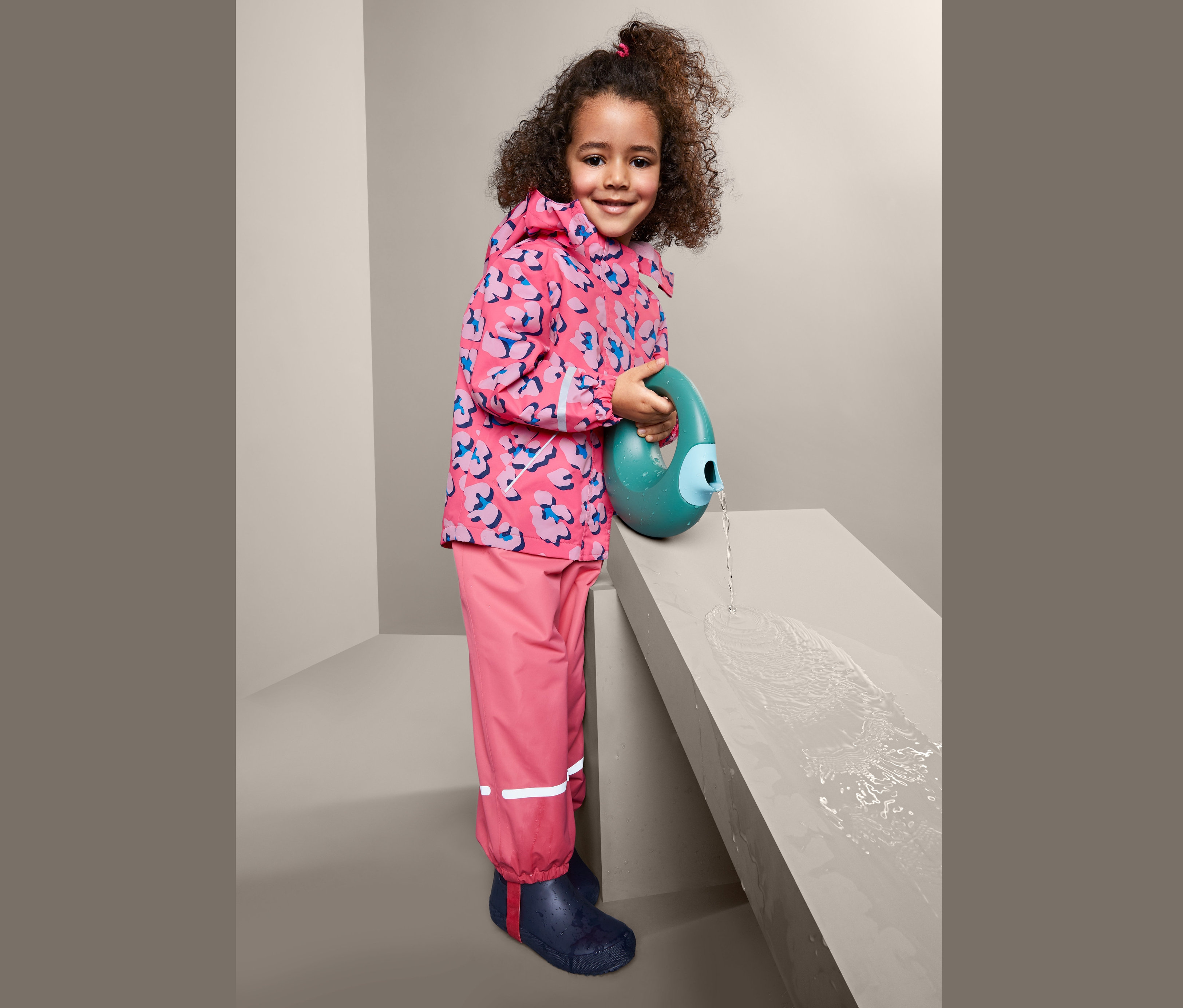 Dívka v termobundě do deště lije vodu z modrozelené konve na betonovou plochu.