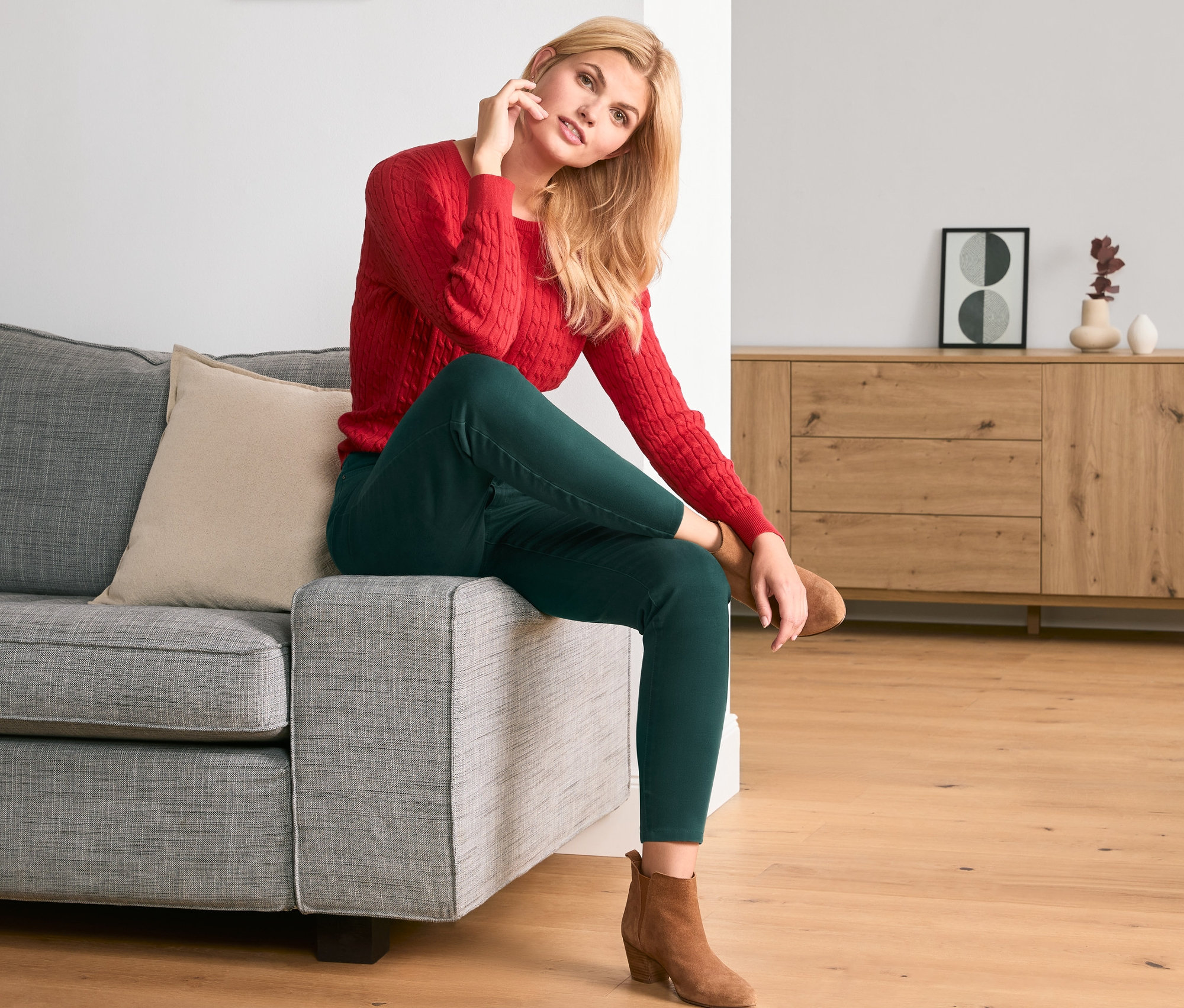 Blondýna sedí na šedé pohovce a má na sobě zelené keprové kalhoty a červený svetr.