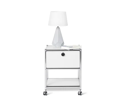 Na bílém pozadí je bílý kovový stolek CN3 se zásuvkou, na kterém stojí lampa a leží kniha.