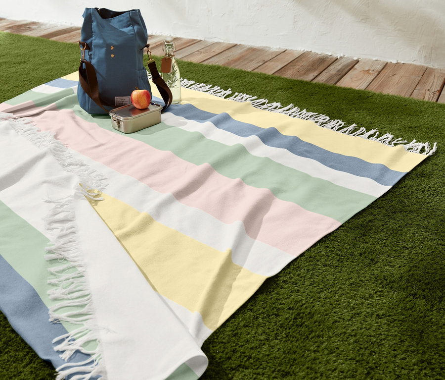 Pruhovaná XL deka na piknik s třásněmi na trávě s taškou, obědovým boxem, jablkem a lahví vody.