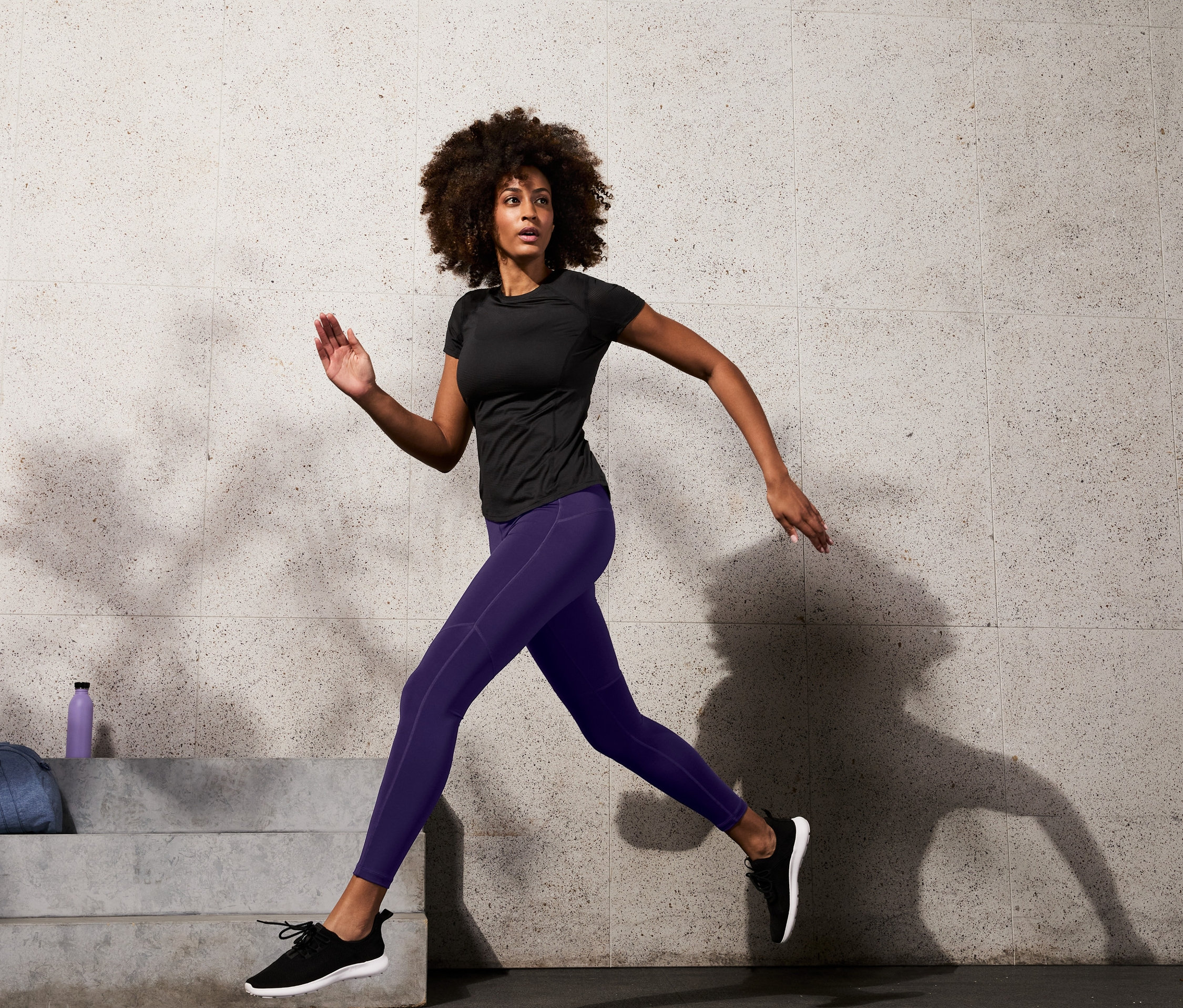 Žena v černém sportovním tričku a stahovacích sportovních legínách běží.