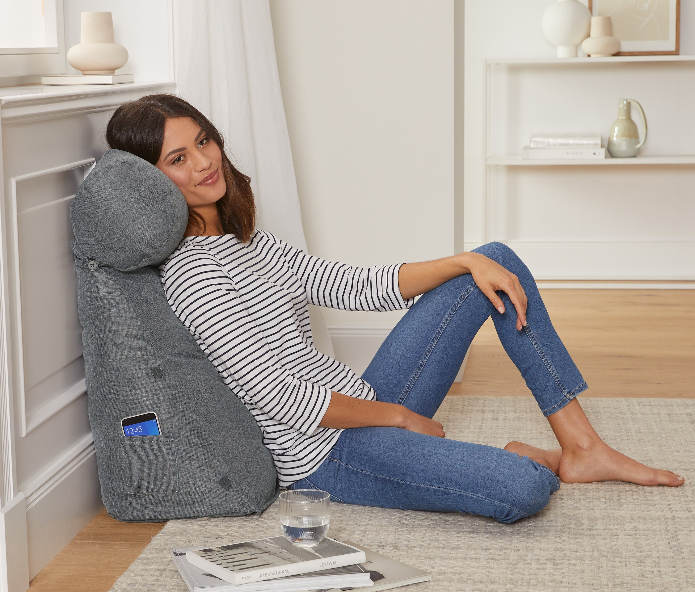 Žena sedí opřená o šedý polštář za záda se šíjovým polštářkem.