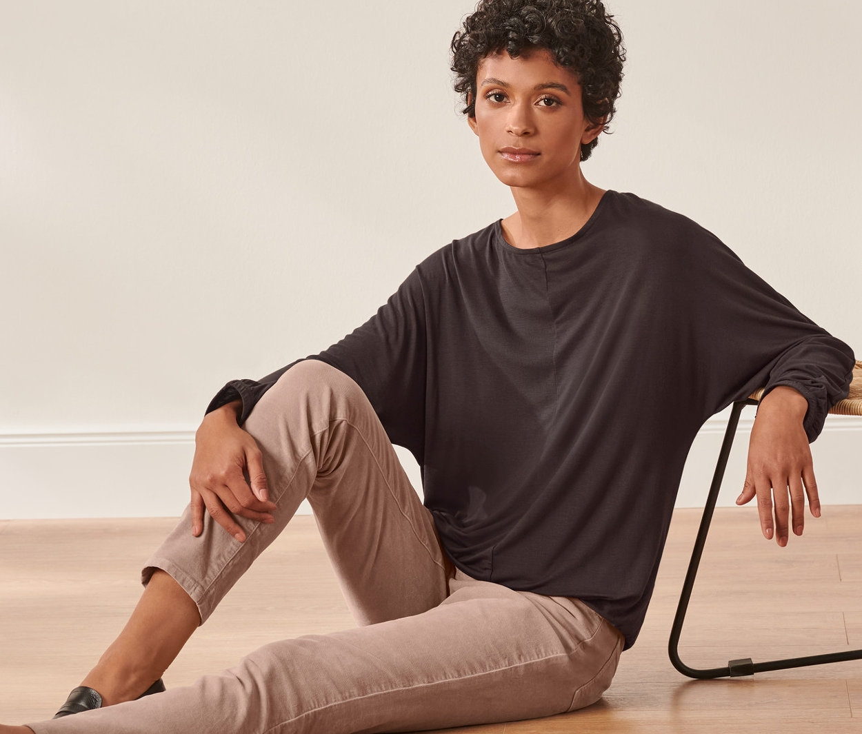 Žena sedí na podlaze vedle proutěného křesla a má na sobě triko s netopýřími rukávy.