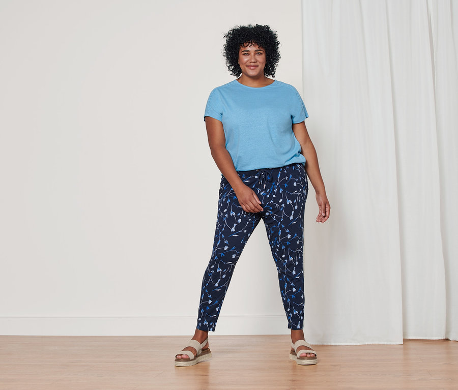 Žena stojí a má na sobě světle modré tričko ze směsi bavlny a lnu, tmavě modré žerzejové kalhoty s celoplošným potiskem a kožené sandály na platformě.