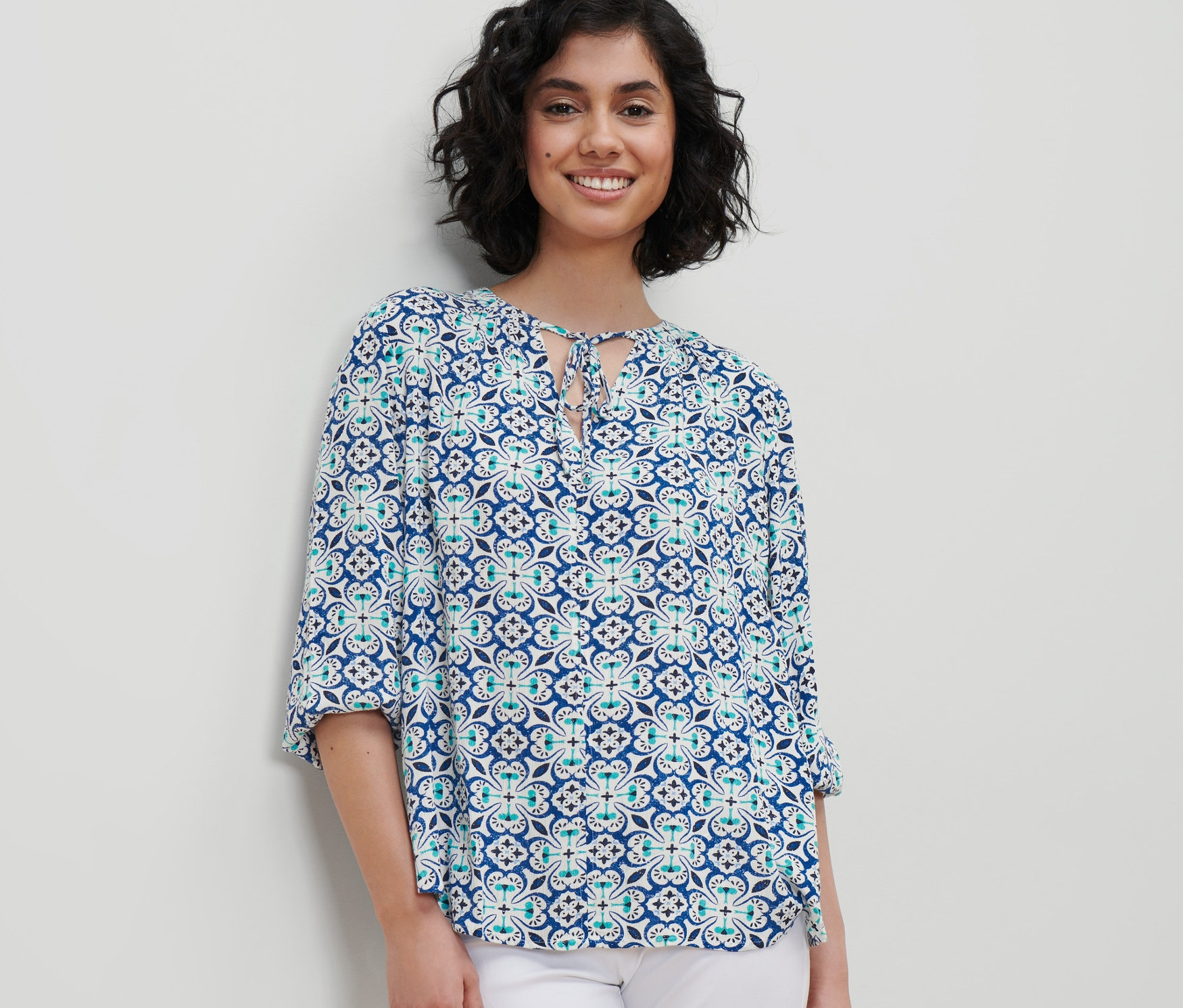 Žena má na sobě tunikovou halenku s celoplošným potiskem a bílé kalhoty.