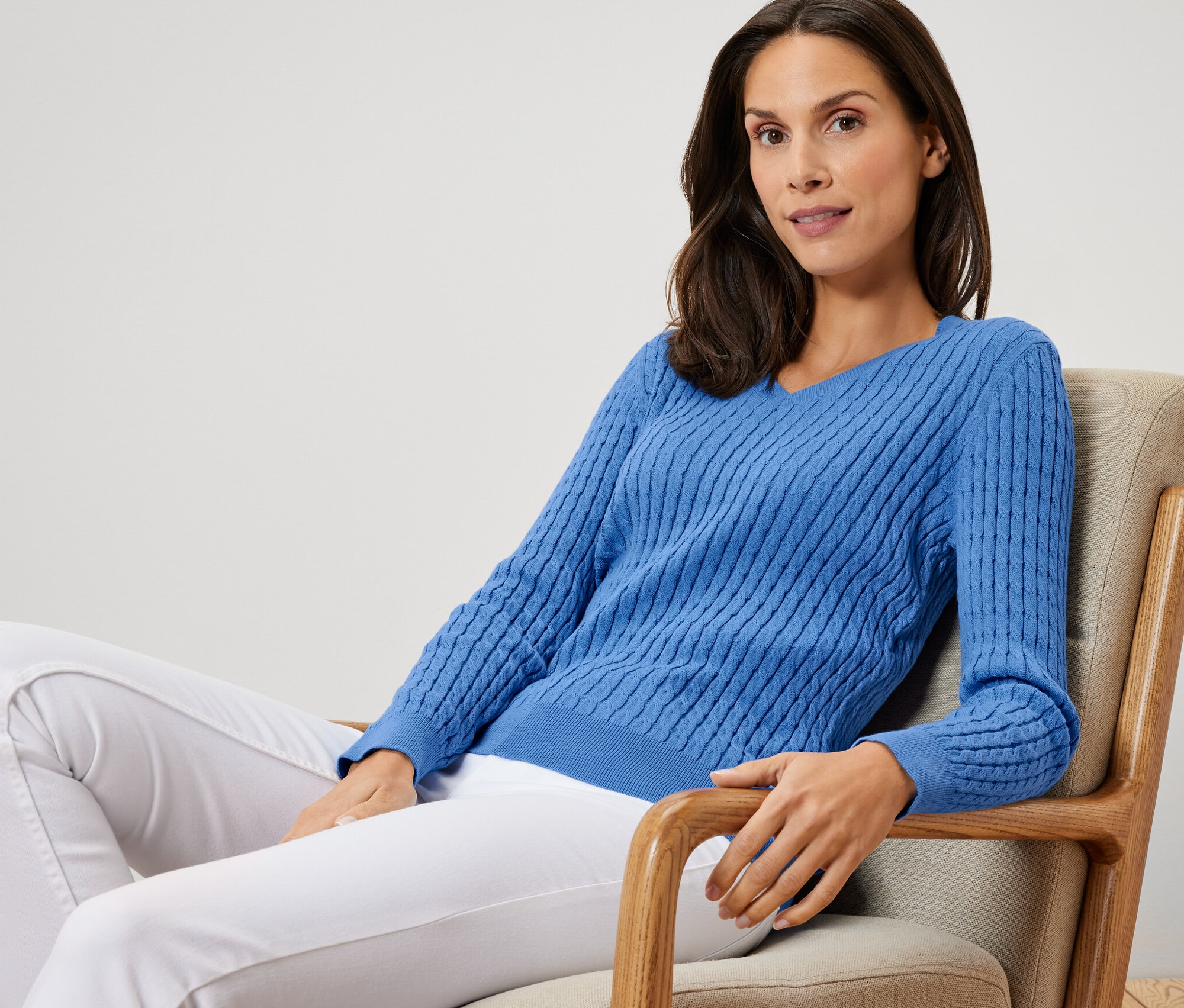 Žena sedí v křesle a má na sobě modrý svetr s copánkovým vzorem.