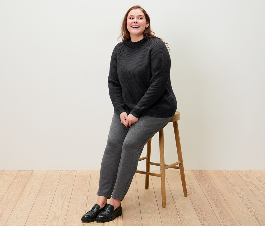 Žena sedí na stoličce a má na sobě černý svetr z hrubého úpletu se stojáčkem a kalhoty joggpant.