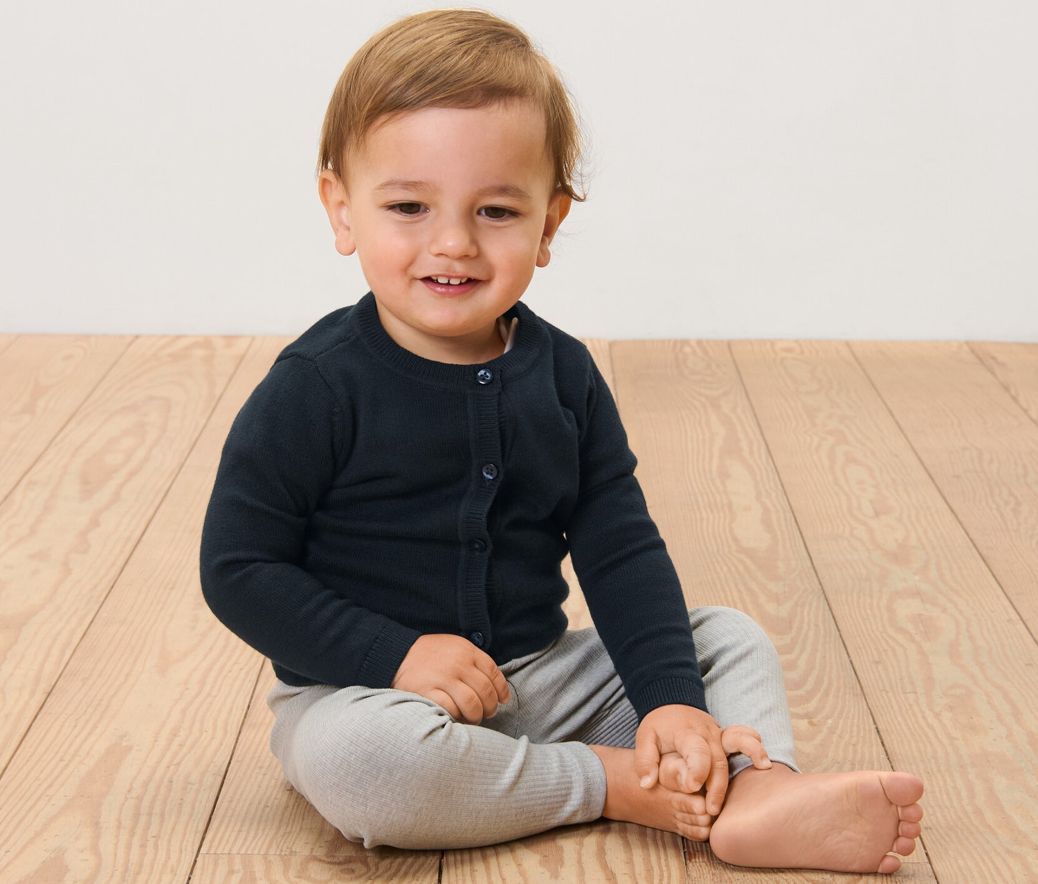 Dítě sedí na dřevěné podlaze a má na sobě dětský pletený svetr.