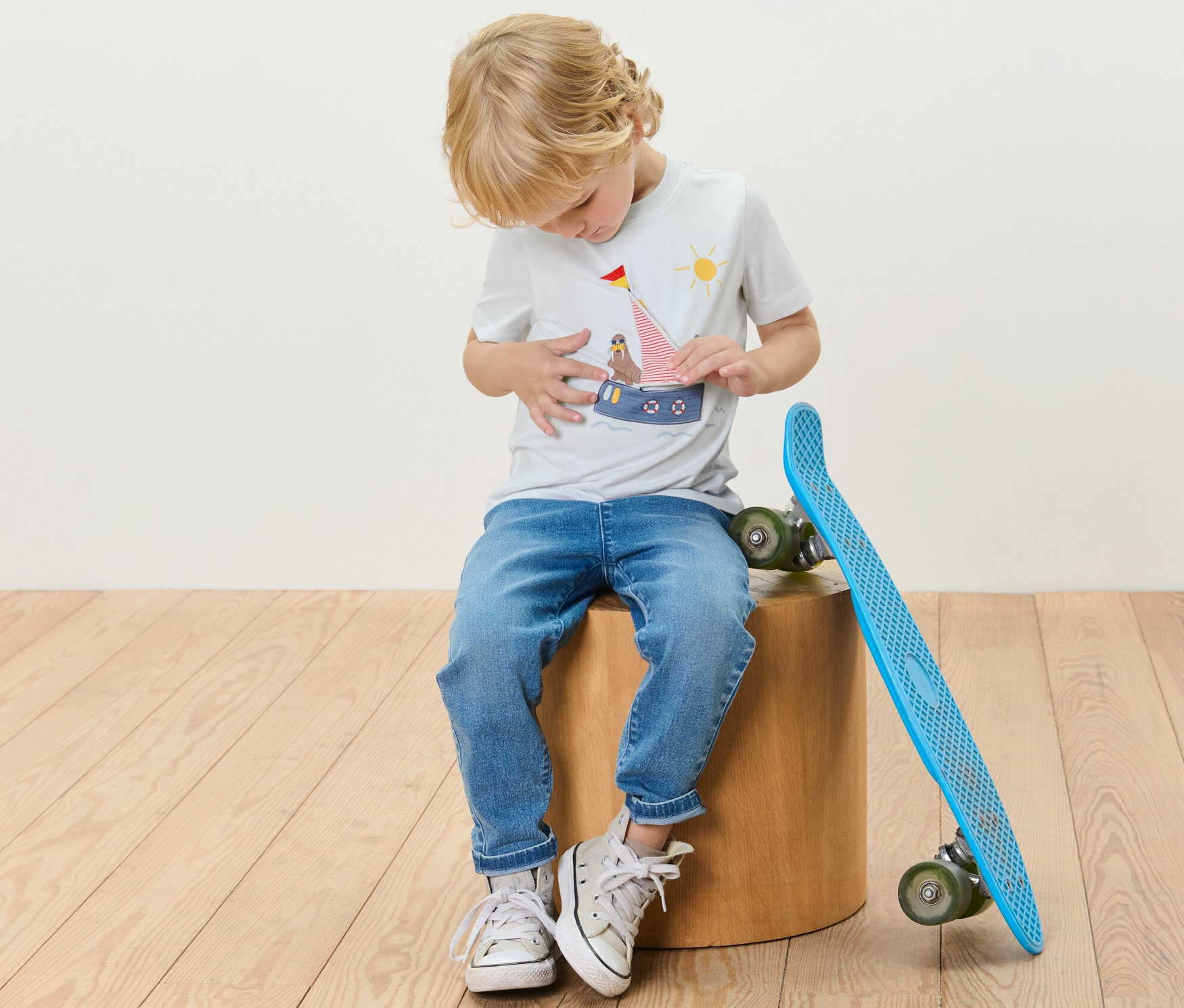 Chlapec sedí na dřevěném špalku vedle modrého skateboardu a má na sobě tričko s námořnickou aplikací.