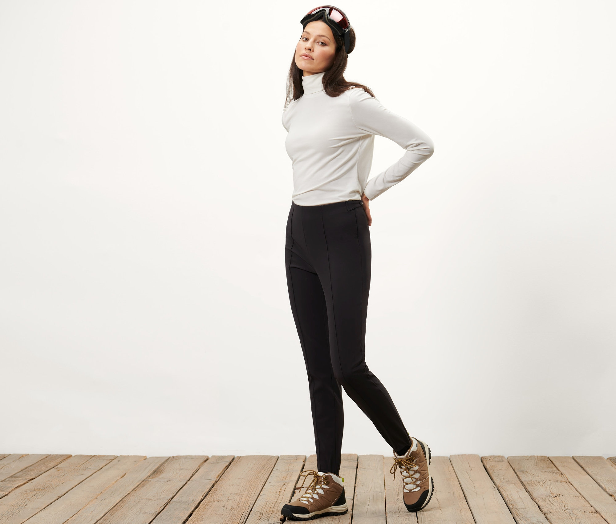 Žena pózuje v softshellových lyžařských legínách, bílém roláku a lyžařských brýlích.