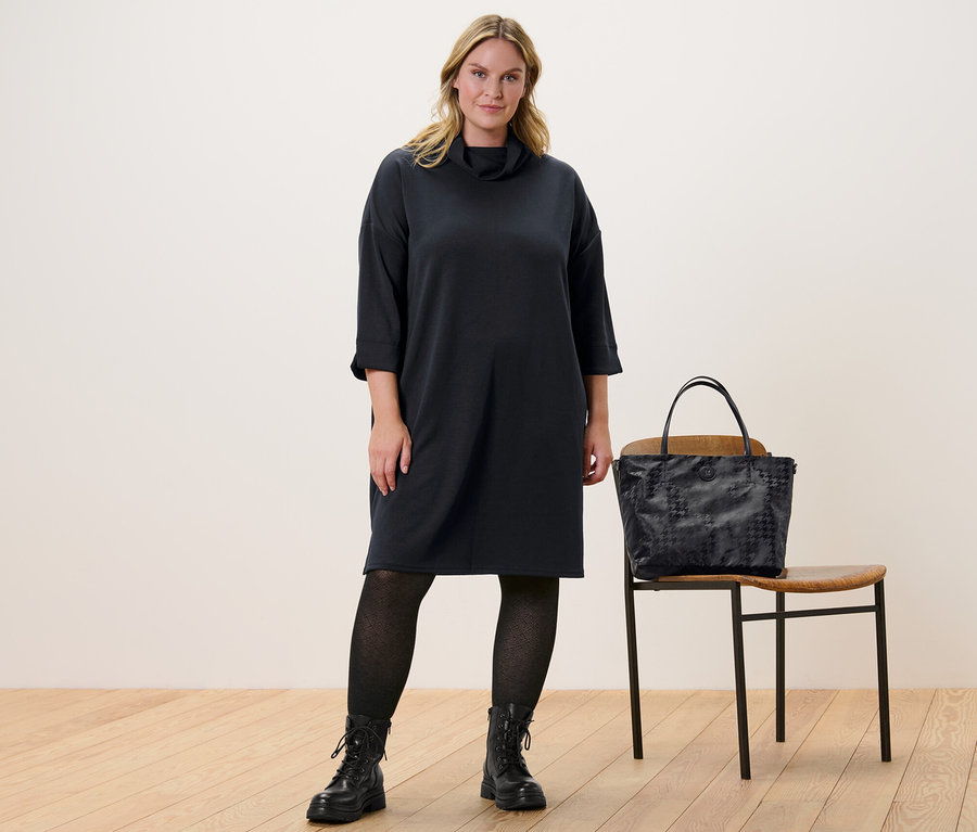 Žena stojí a má na sobě teplákové šaty z modalové směsi a strukturované pletené punčocháče. Vedle ní stojí židle a na ní je kabelka.