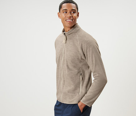 Muž pózuje v béžové fleecové bundě a tmavě modrých kalhotách.